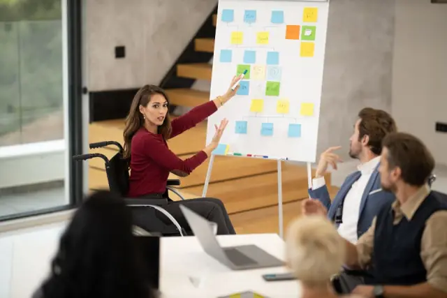 Une femme en fauteuil roulant anime un atelier de projet devant un tableau de post-it